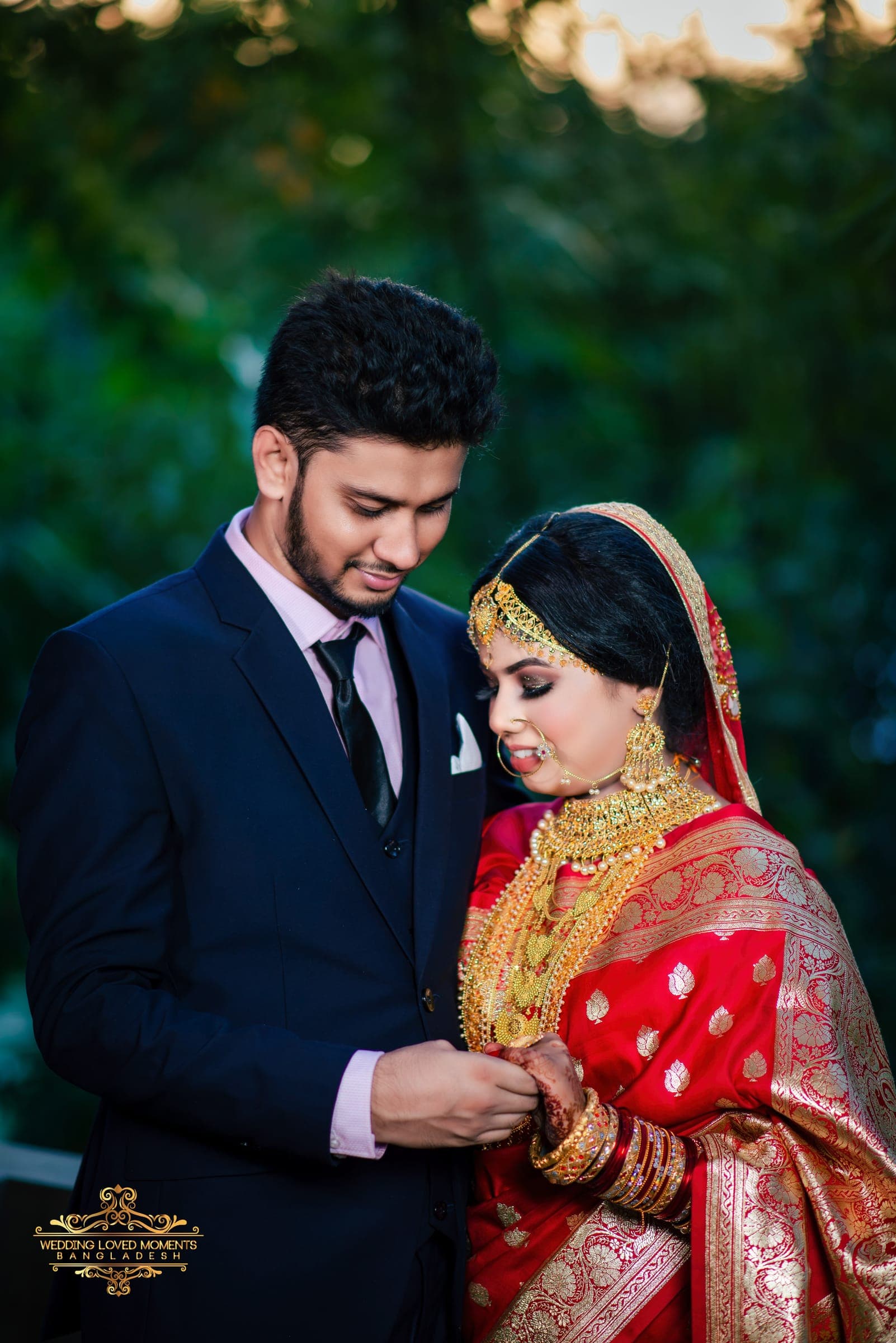 Groom in a navy suit holding the bride's hand as she wears a red Banarasi saree, framed by garden greenery
