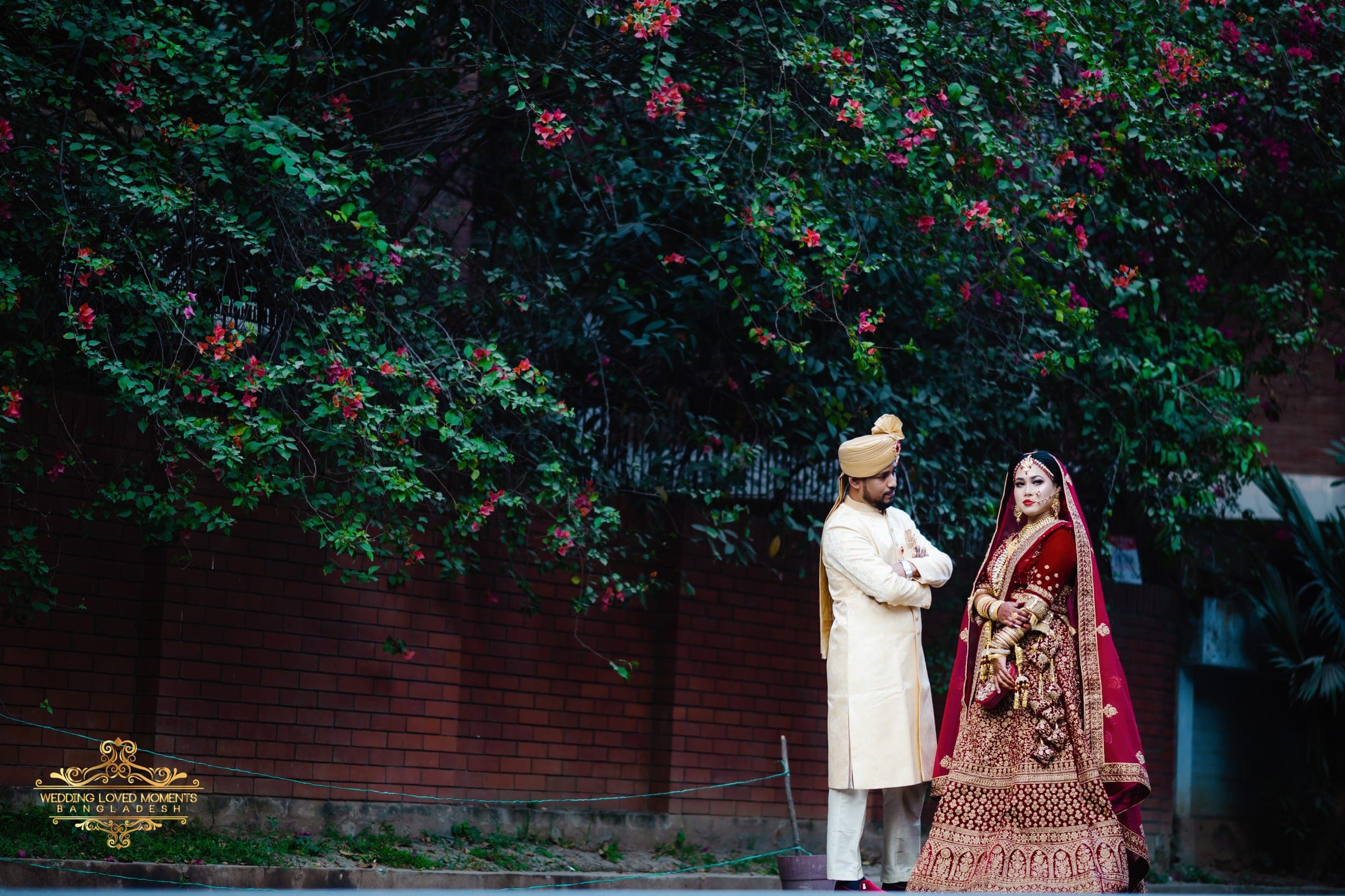 Bride and groom standing beside a bougainvillea-covered wall, she in a red lehenga and he in an ivory sherwani
