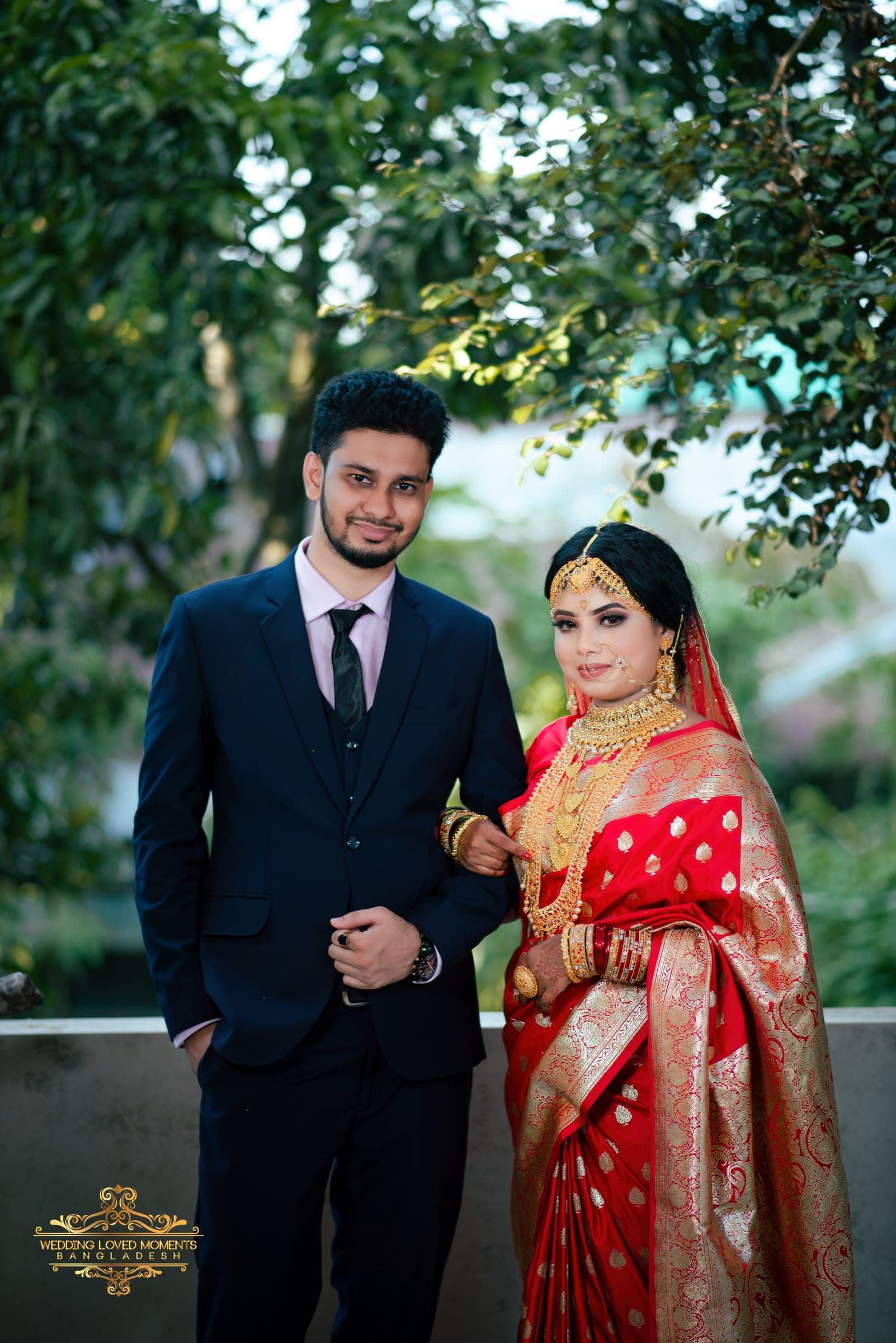 Couple standing beneath trees, the bride in a red saree and the groom in a navy suit