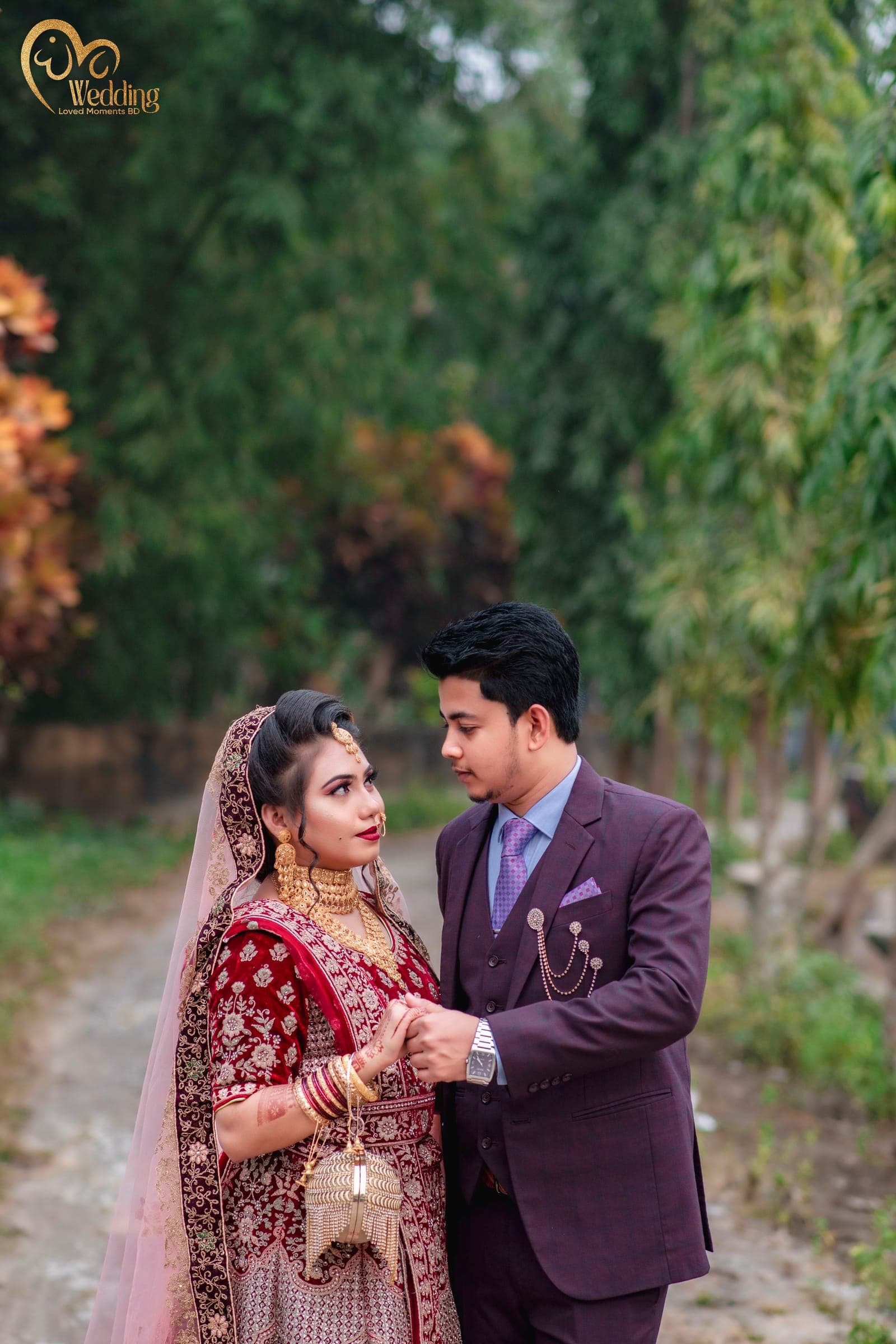 Couple walking hand in hand down a tree-lined path in maroon wedding attire