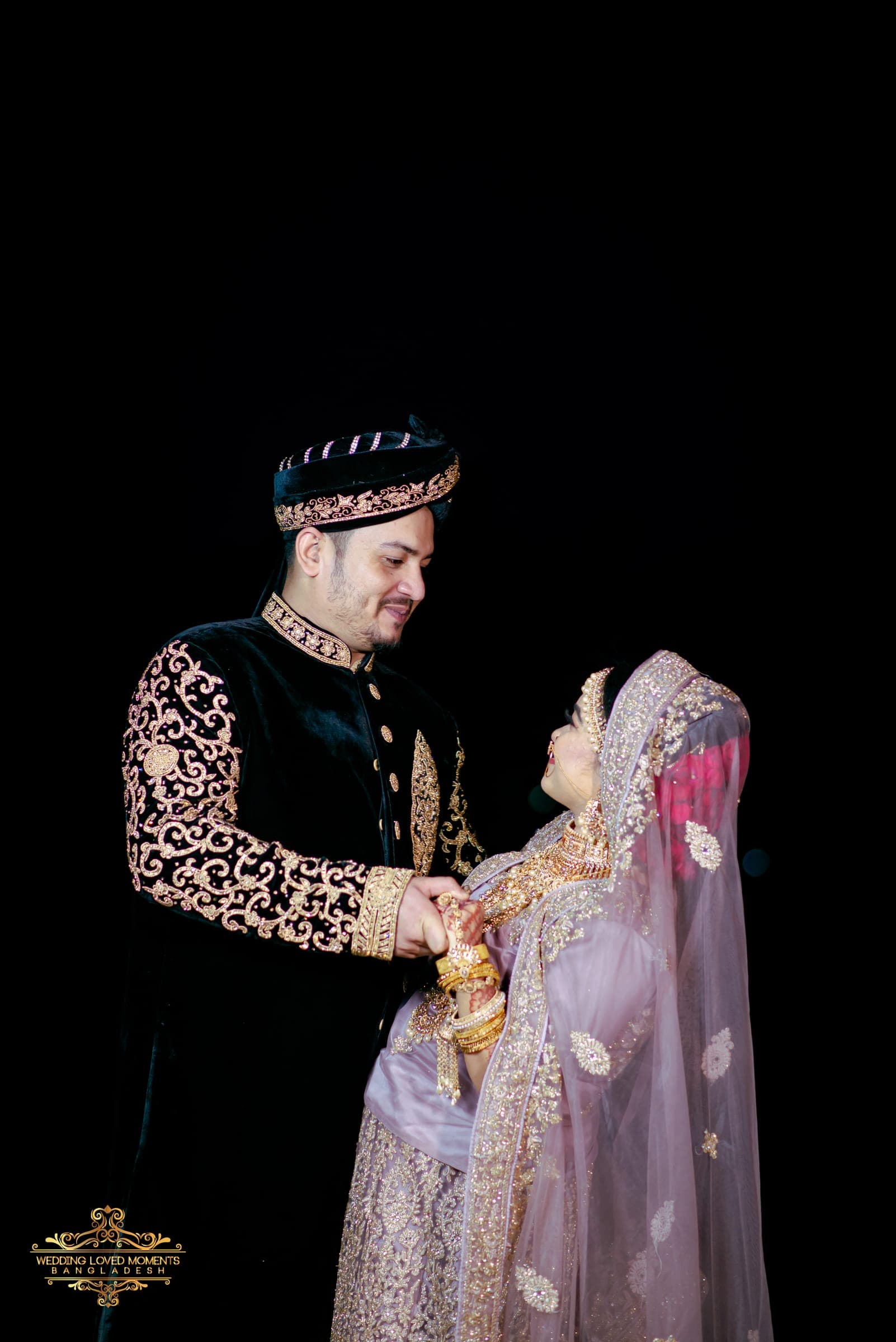 Couple in a black velvet sherwani and lilac bridal outfit against a dark backdrop