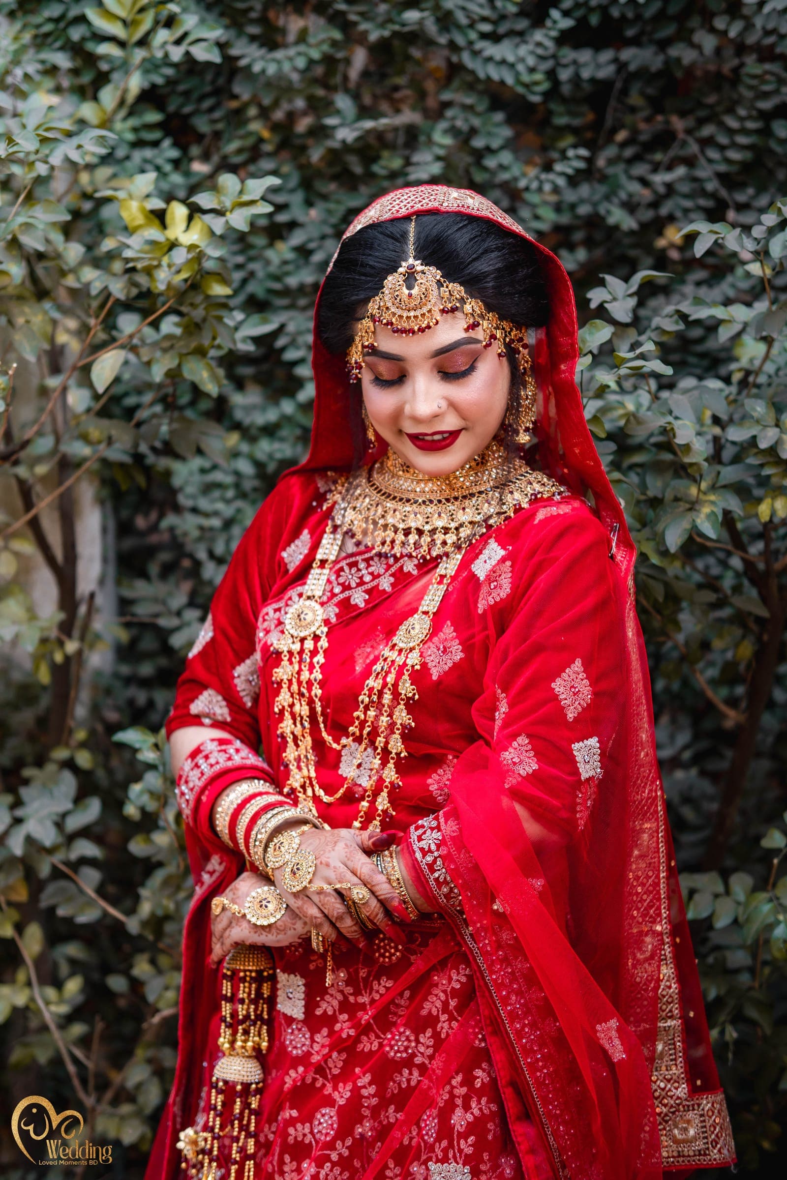 Bride in a red saree and layered gold jewellery looking down softly among green leaves