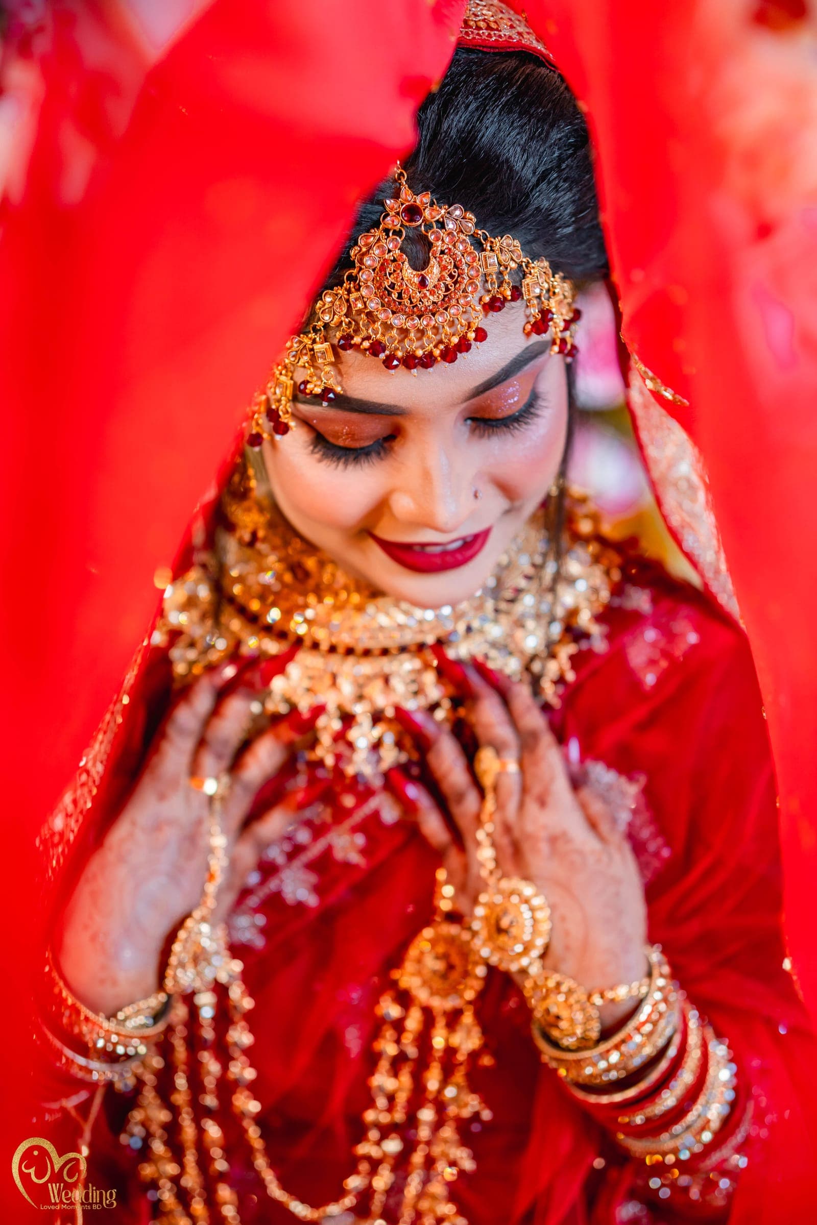 Close portrait of a smiling bride beneath a red veil, hands adorned with gold jewellery
