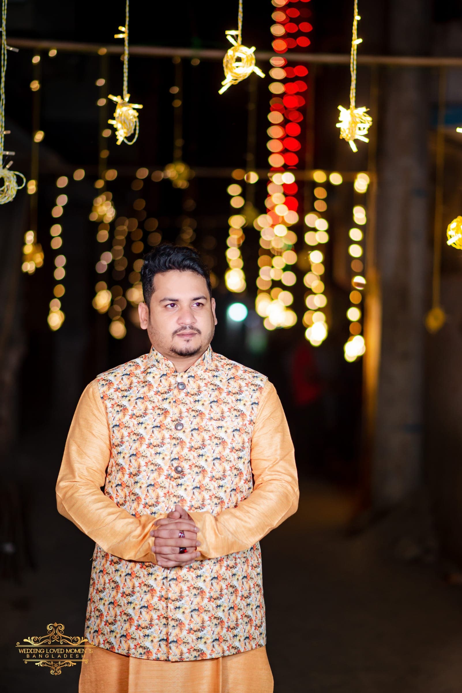 Groom in a peach panjabi and paisley waistcoat under hanging fairy lights