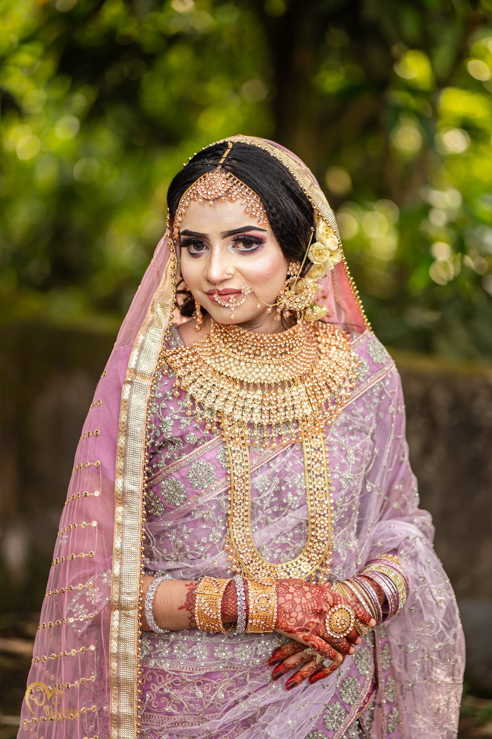 Bride in a lilac lehenga with heavy gold jewellery in a leafy outdoor setting